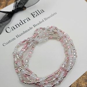 May include: Handmade beaded bracelets in shades of pink, silver, and clear beads. The bracelets are arranged on a white surface with the text "Candra Ella Custom Handmade Beaded Bracelets". A black ribbon is in the background.