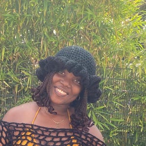 Pode incluir: Uma pessoa usando um chapéu de crochê preto com uma borda franjada e um top de crochê preto. A pessoa está sorrindo e olhando para a câmera.