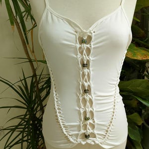 Può includere: Top bianco con spalline sottili e dettagli in macramè sul davanti e sui lati. Il macramè è impreziosito da piccole perline decorative. Il top è realizzato in un materiale morbido ed elastico.