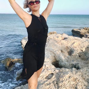May include: A black sleeveless dress with a unique design, featuring a lace-up neckline and a side slit. The dress is worn by a person standing on a rocky shoreline with the ocean in the background. The person is wearing sunglasses.