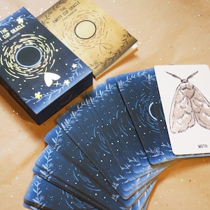 May include: A deck of oracle cards titled "The Empty Cup Oracle" with a matching box and guidebook. The cards and box feature a dark blue background with white floral and celestial designs. One card shows a moth illustration.