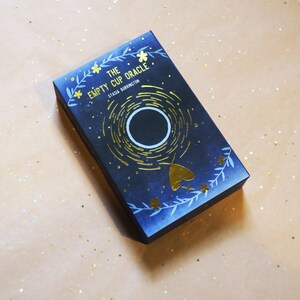 May include: A dark blue oracle card box with gold foil accents. The box features the title "THE EMPTY CUP ORACLE" and the author's name, STASIA BURRINGTON. The design includes a central circular element, a moth, and floral details.