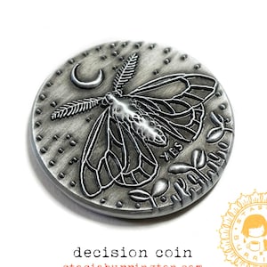 May include: A silver-colored decision coin with a moth design, a crescent moon, and the words "YES" and "Maybe". The coin has a textured border and is set against a white background. The text "decision coin" and a website address are also visible.