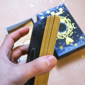 May include: A deck of tarot cards with gold gilded edges, held in a hand. The cards are black with gold designs, and the box is navy blue with gold accents. The cards are approximately 5 inches long.