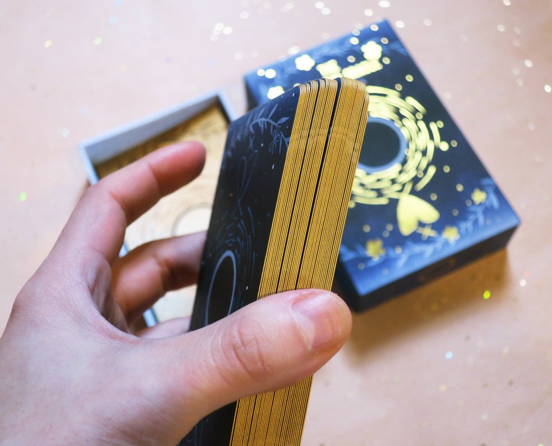 May include: A deck of tarot cards with gold gilded edges, held in a hand. The cards are black with gold designs, and the box is navy blue with gold accents. The cards are approximately 5 inches long.