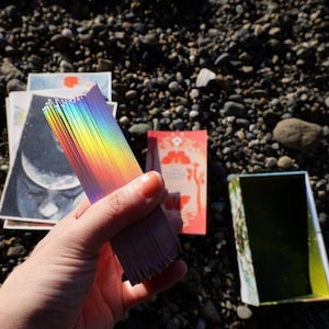 May include: A deck of rectangular cards with holographic rainbow edges held in a stack. The cards feature various illustrations, including a moon design. An open rectangular box with a green interior sits nearby on a dark, pebbled surface.
