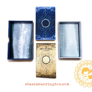 May include: An overhead shot of an oracle card set. The set includes a box with the words "empty cup oracle" on the side, and the text "THE EMPTY CUP ORACLE" on the front of the card box. The cards and boxes are blue and beige.