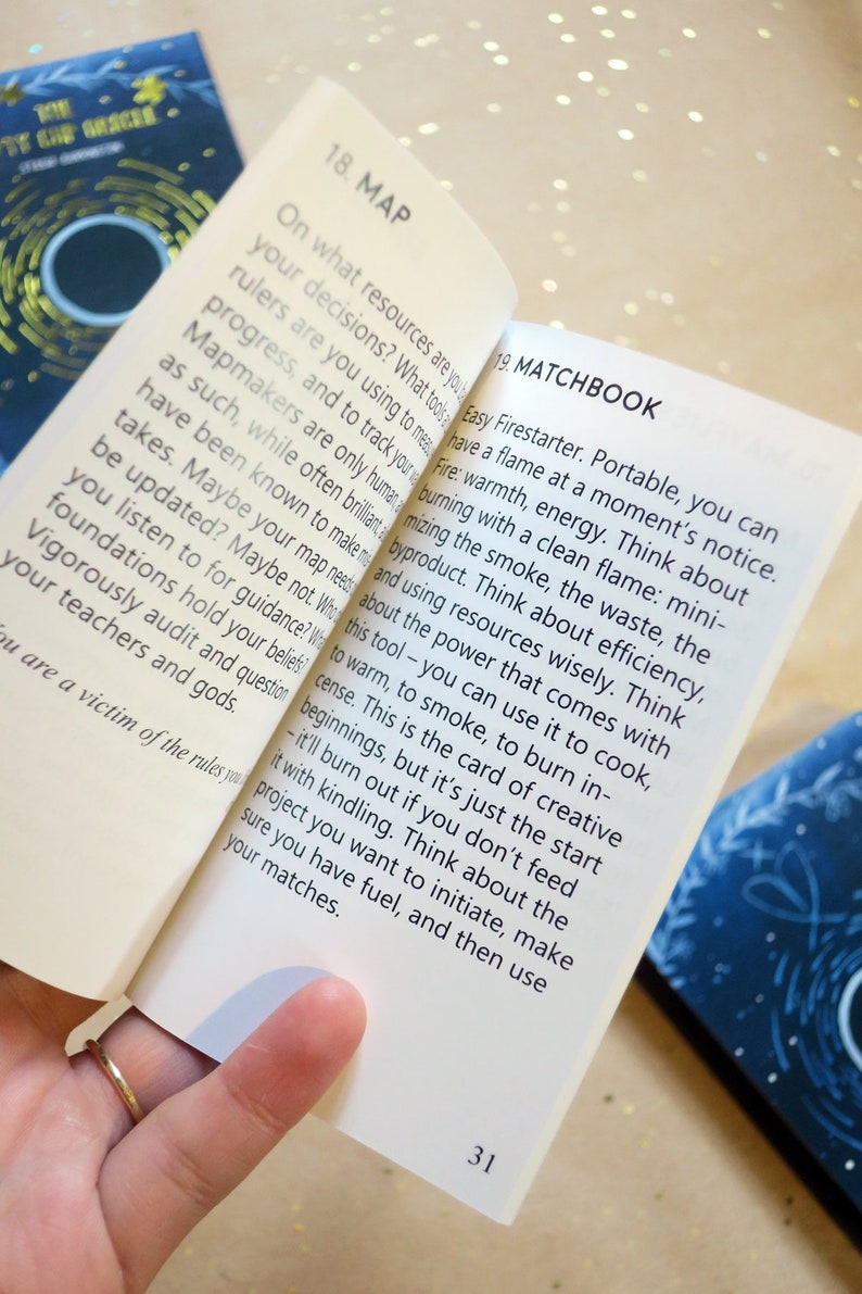 May include: Open pages of a book titled "The City of Oracle" with text about "Map" and "Matchbook". The pages are held open by a hand with a gold ring. The background is a light, glittery surface.