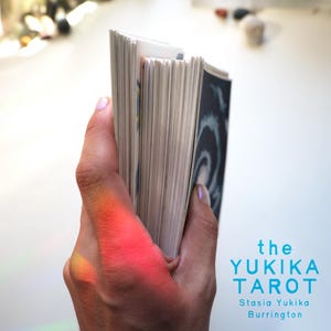 May include: A deck of tarot cards held in a vertical stack, featuring white edges and a dark card back with a swirling design. The text on the right side reads: the YUKIKA TAROT, Stasia Yukika Burrington. A bright red light reflection is visible.