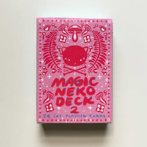 May include: A pink playing card box with a red cat illustration and the words "MAGIC NEKO DECK 2". The box features a repeating pattern of mushrooms, flowers, and ferns. The text "54 CAT PLAYING CARDS" is also visible.