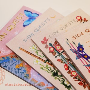 May include: A collection of illustrated notepads with the words "Main Quest" and "Side Quests" in gold lettering. Each notepad features a different design, including a butterfly, a wand, a sword, and arrows, with floral accents. The notepads are in shades of pink and beige.