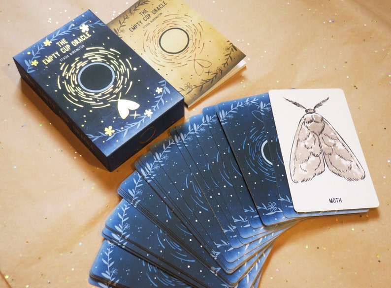 May include: A deck of oracle cards titled "The Empty Cup Oracle" with a matching box and guidebook. The cards and box feature a dark blue background with white floral and celestial designs. One card shows a moth illustration.