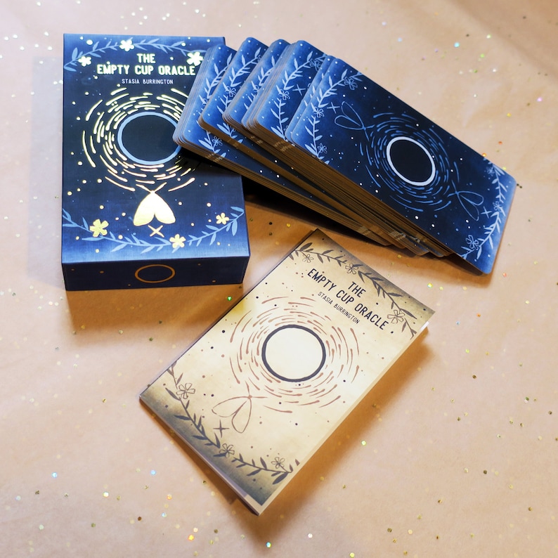 May include: An oracle deck titled "The Empty Cup Oracle" by Stasia Burrington. The deck features a navy blue box and card backs with a celestial design. The cards have a circular design with floral and moth illustrations.