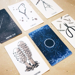May include: A collection of illustrated cards featuring various designs. The cards display images of a telescope, a slingshot, a shooting star, scissors, mushrooms, and a celestial design. The cards are arranged on a light brown surface with gold glitter.