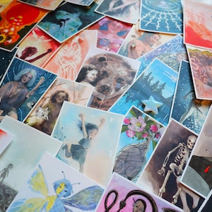May include: A collection of rectangular art cards featuring diverse watercolor illustrations, including a skull, a bear, a skeleton, and a volcanic eruption. One card displays the text 'the YUKIKA TAROT Stasia Yukika Burrington' in teal.