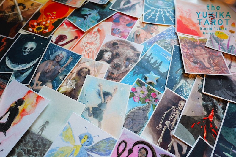May include: A collection of rectangular art cards featuring diverse watercolor illustrations, including a skull, a bear, a skeleton, and a volcanic eruption. One card displays the text 'the YUKIKA TAROT Stasia Yukika Burrington' in teal.