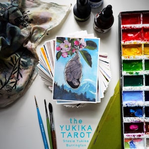 May include: A stack of tarot cards titled the YUKIKA TAROT by Stasia Yukika Burrington, featuring a top card with a cocoon illustration under pink blossoms. Includes a watercolor paint set, three paintbrushes, and three ink bottles.