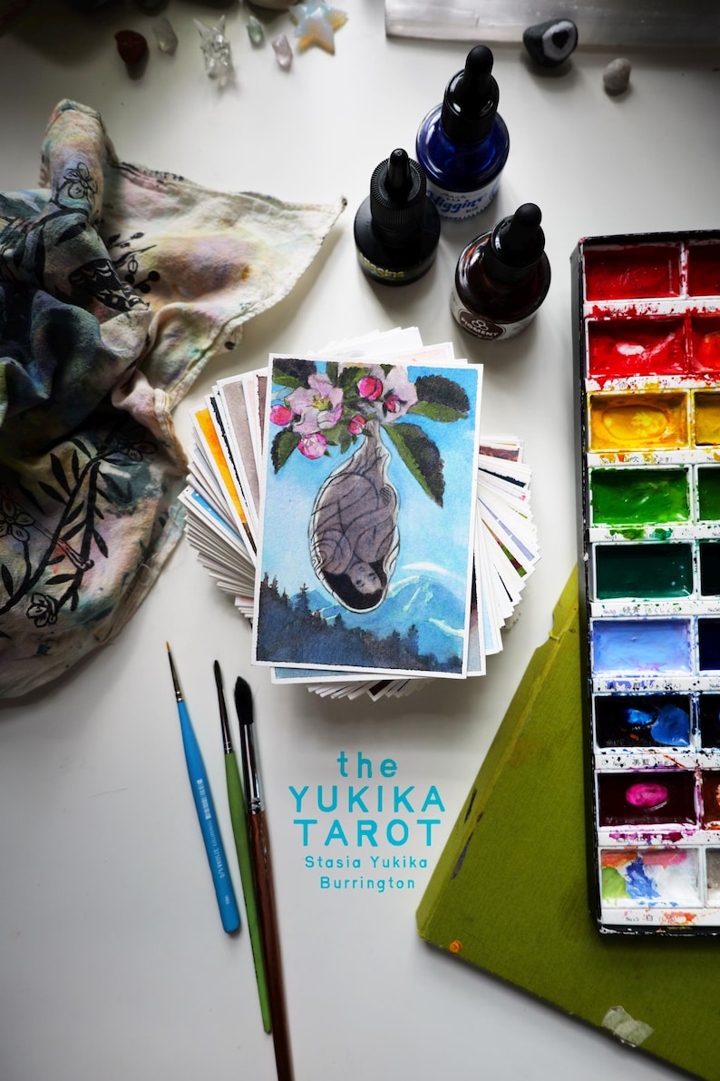 May include: A stack of tarot cards titled the YUKIKA TAROT by Stasia Yukika Burrington, featuring a top card with a cocoon illustration under pink blossoms. Includes a watercolor paint set, three paintbrushes, and three ink bottles.