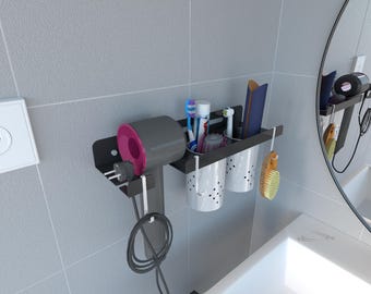 May include: A black wall-mounted bathroom organizer with a hairdryer, two white cups, and a toothbrush holder. The hairdryer is gray and pink. The organizer also holds toothbrushes, toothpaste, and a brush. The organizer is mounted on a gray tiled wall.