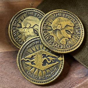 Könnte beinhalten: Drei antike, goldfarbene Münzen mit geprägten Designs und Text, darunter "THE EMPIRE IS LAW" und "THE LAW IS SACRED". Die Münzen sind auf einem Holztablett angeordnet, mit einem Keramikobjekt und einem dunkelgrünen Stoff im Hintergrund.