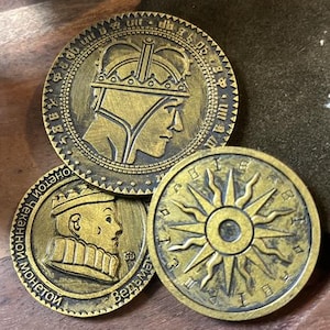 May include: Three antique-style gold coins with intricate designs. One coin shows a crowned profile, another a sunburst, and the third a profile with text. The coins are displayed on a wooden surface.