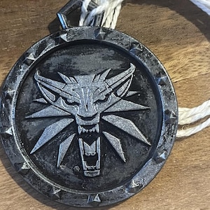 Puede incluir: Un colgante circular plateado con un emblema de cabeza de lobo y un cordón blanco. El colgante tiene una superficie texturizada y un diseño detallado. El cordón está hecho de hilos blancos trenzados. El colgante es probablemente una joya o un objeto de colección.