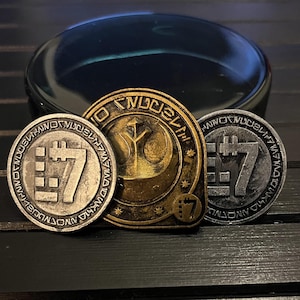 May include: Three metallic coins with intricate designs. The central coin is gold-colored, featuring a symbol and text. Two silver-colored coins flank it, each with a stylized number 7 and surrounding text. The coins are set against a dark background.
