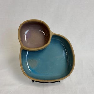 Puede incluir: Una fuente de cerámica hecha a mano con dos secciones conectadas. La sección más grande es azul brillante, mientras que la más pequeña es morada apagada. El borde es de color arcilla natural sin esmaltar. El plato se exhibe sobre un soporte de metal negro.