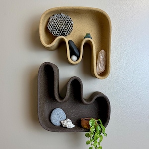 Op de afbeelding: Twee abstract gevormde wandplanken in beige en bruin. De beige plank bevat een honingraat, stenen en een kristal. De bruine plank toont een kleine plant, een zeeschelp en een steen. Decoratieve woondecoratie.