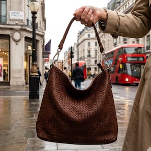 May include: A brown woven leather shoulder bag with a long strap and silver-tone hardware. The bag is held up in front of a street scene with a red double-decker bus in the background.
