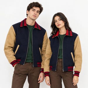 May include: A varsity-style jacket featuring a navy blue wool-blend body, tan sleeves, and a red and navy striped collar and cuffs. The jacket has a front zip closure, two side pockets with tan trim, and is worn over a dark green shirt.