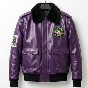 Könnte beinhalten: Eine lila Lederjacke mit einem schwarzen Kunstfellkragen. Die Jacke hat zwei Vordertaschen, einen schwarzen gerippten Saum und Bündchen sowie einen Joker-Aufnäher auf der Brust. Ein Spielkarten-Aufnäher befindet sich auf dem linken Ärmel.