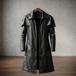 Pode incluir: Um casaco longo de couro preto com um detalhe de ombro tipo capa pendurado em um cabide de madeira. O casaco tem um design clássico e sob medida com gola e uma silhueta longa e fluida. O couro parece desgastado, dando-lhe um visual vintage.