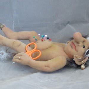 May include: A handmade troll doll with a large nose, white hair, and a smiling expression. The doll has colourful pins stuck in its chest and is holding orange scissors. A thimble is on one finger. The doll is lying down.