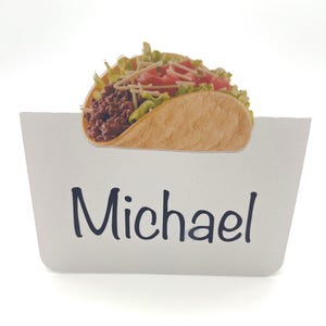 May include: A white place card with the name "Michael" in black script. A detailed taco illustration sits atop the card, featuring a golden-brown tortilla filled with seasoned ground meat, shredded lettuce, tomatoes, and cheese.