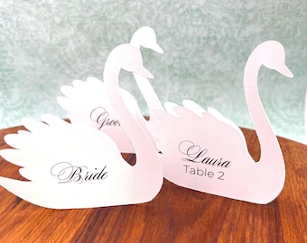 May include: Three pale pink swan-shaped paper place cards with die-cut wing details. The cards feature black printed text including the words Bride, Groom, Laura, and Table 2 in a decorative script and sans-serif font, standing upright.