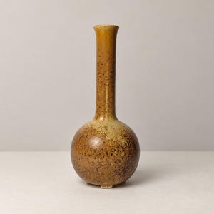 May include: A ceramic bud vase with a spherical base and a tall, slender neck. The surface features a mottled, speckled glaze in shades of earthy brown, tan, and ochre, with a smooth, glossy finish throughout the entire vessel.