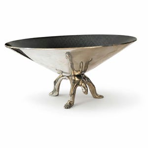Peut inclure: Un bol décoratif avec un extérieur argenté lisse et poli et un intérieur noir mat texturé. Le bol est soutenu par trois pieds métalliques en forme de silhouettes humaines en position accroupie, offrant une base stable au récipient.