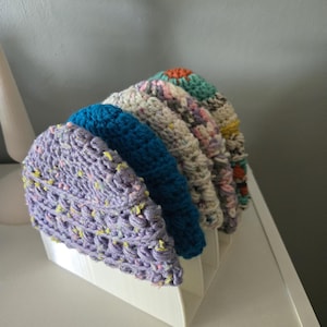 May include: A display of five colorful, handmade crochet hats. The hats are in shades of purple, blue, white, and multicolored patterns. They are arranged in a white, angled display holder, showcasing the variety of designs.