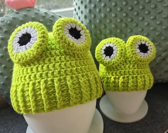 May include: Two lime green crochet frog hats with white and black eye details sit on white egg-shaped stands. The hats are displayed on a grey surface, with a cactus-shaped pillow in the background.