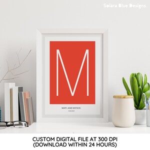 Könnte beinhalten: Gerahmter Druck mit einem weißen Buchstaben "M" auf rotem Hintergrund. Der Druck enthält den Namen "MARY JANE WATSON" und das Datum "12.02.2021". Das Kunstwerk steht auf einem weißen Regal mit dekorativen Gegenständen.
