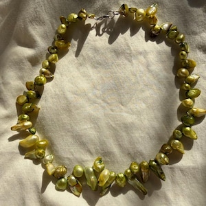 May include: A necklace composed of numerous irregular, organic-shaped pearls in varying shades of lime green, olive, and chartreuse. The pearls are strung together in a single strand, secured with a small, polished silver-toned metal clasp.