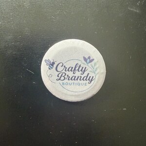 May include: A circular pin-back button with a white fabric-like surface. The design features a small blue bee, a purple flower, and the dark blue text "Crafty Brandy BOUTIQUE" centered within a thin circular line. The word handmade is visible.