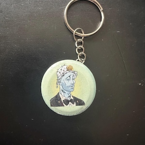 May include: A circular button keychain featuring a pale blue-skinned character wearing a white patterned bucket hat, a black suit jacket, and a black bow tie. The background of the button is a soft yellow, and the text reads Boutique Handmade.