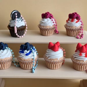 May include: Miniature cupcakes with diverse toppings, such as an Oreo, raspberries, blueberries, and red bows. Each cupcake features white frosting and a beaded chain. The cupcakes are presented on a light-coloured shelf, showcasing a range of designs.