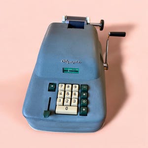 Puede incluir: Una calculadora mecánica Olympia vintage en un tono azul-gris apagado. La calculadora presenta un teclado numérico completo con teclas verdes y blancas, una manivela y un rollo de papel. La palabra "Olympia" está impresa en la parte delantera.