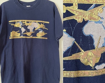 Puede incluir: Una camiseta azul marino de manga corta con un estampado gráfico dorado de varios aviones militares, un tanque y un submarino sobre un mapa mundial azul claro. El texto "GENERAL DYNAMICS" aparece en dorado dentro de barras horizontales.