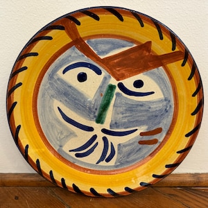 May include: Hand-painted ceramic plate with a whimsical face design. The plate features a yellow border with black accents, a blue and white face, and brown, green, and blue details. A decorative piece for kitchen or dining.