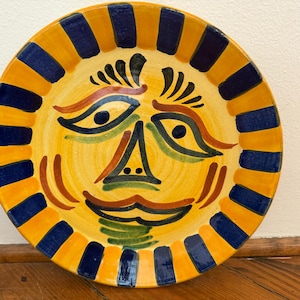 May include: A hand-painted ceramic plate with a sun face design. The plate features a yellow center with a smiling face in shades of orange, green, and black. The rim is yellow with alternating blue and orange rectangular shapes.