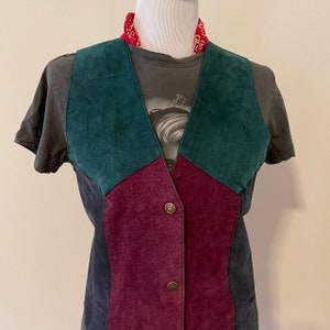 May include: A multi-colored suede vest with a patchwork design. The vest features panels of teal, burgundy, and dark gray suede, fastened with two brass-colored buttons. A red bandana is around the neck.